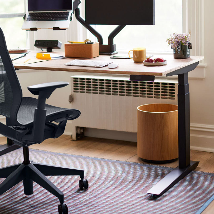 Entorno de oficina en casa con vista parcial de una silla de oficina Cosm con respaldo alto en color noche delante de un escritorio sit-stand Jarvis con patas negras y superficie de madera.