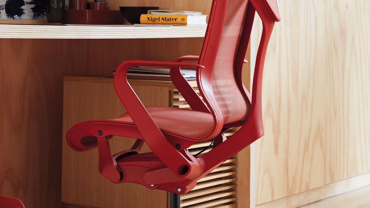 Una silla de oficina Cosm roja de Herman Miller, colocada debajo de un escritorio. Está situada en un ambiente interior con suelos y paredes de madera noble, con un enfoque en el vibrante color de la silla en medio del mobiliario circundante.