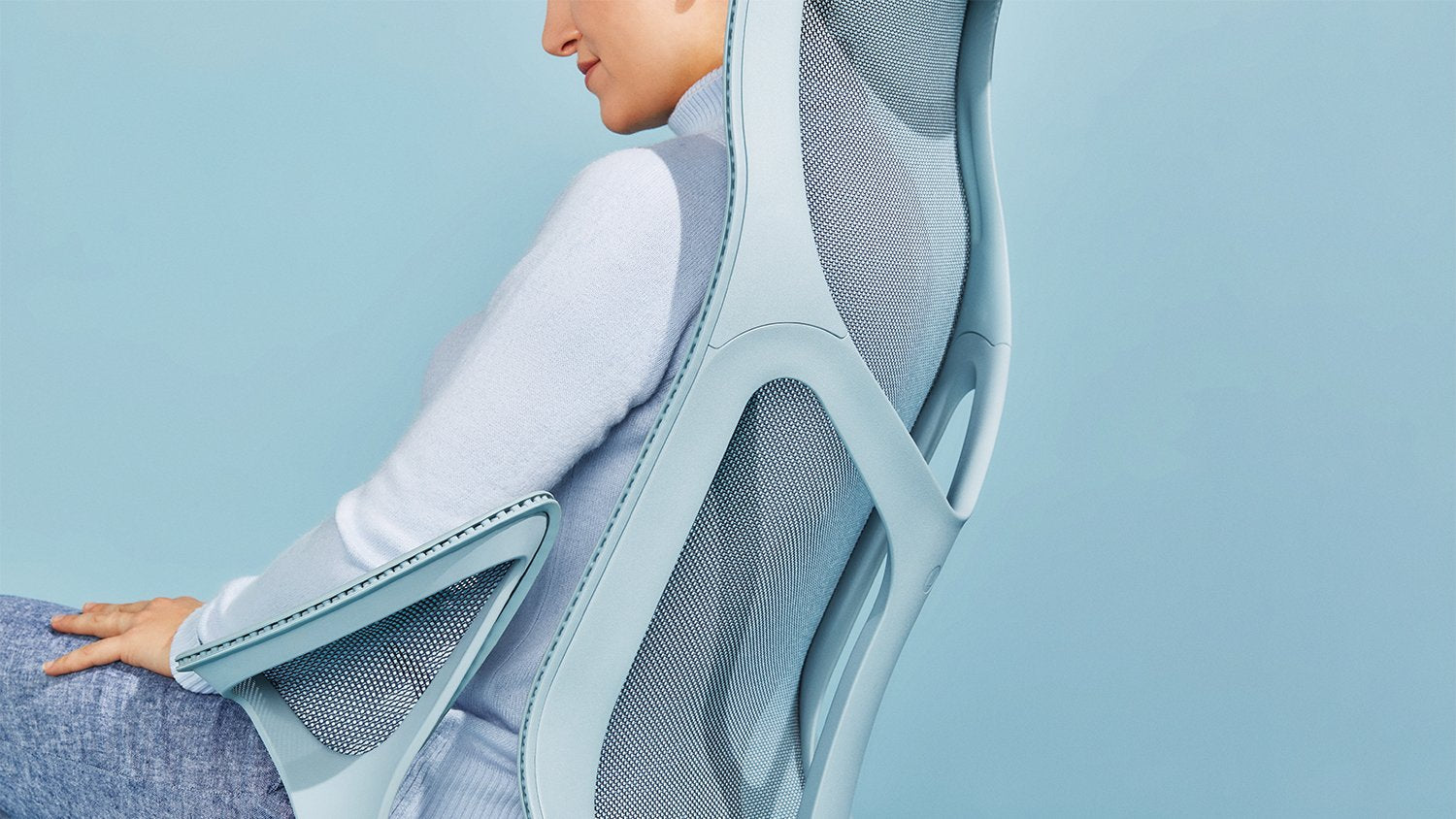 Una persona sentada en una silla de oficina Cosm azul mineral de Herman Miller, vestida con un jersey azul y vaqueros, frente a un fondo liso azul claro.