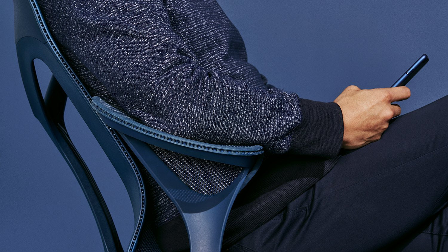Una persona sentada en una silla de oficina Cosm azul anochecer de Herman Miller. Lleva un teléfono en la mano y viste ropa de color azul oscuro sobre un fondo liso azul oscuro.