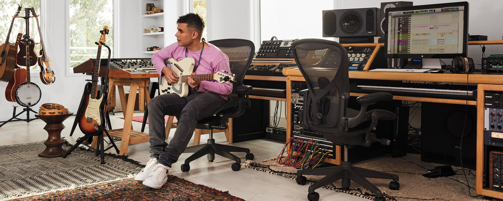 Rostam Batmanglij, músico, productor y compositor, en su estudio musical tocando una guitarra Italia Modena Electric Sitar sentado en una silla de oficina Aeron en grafito.