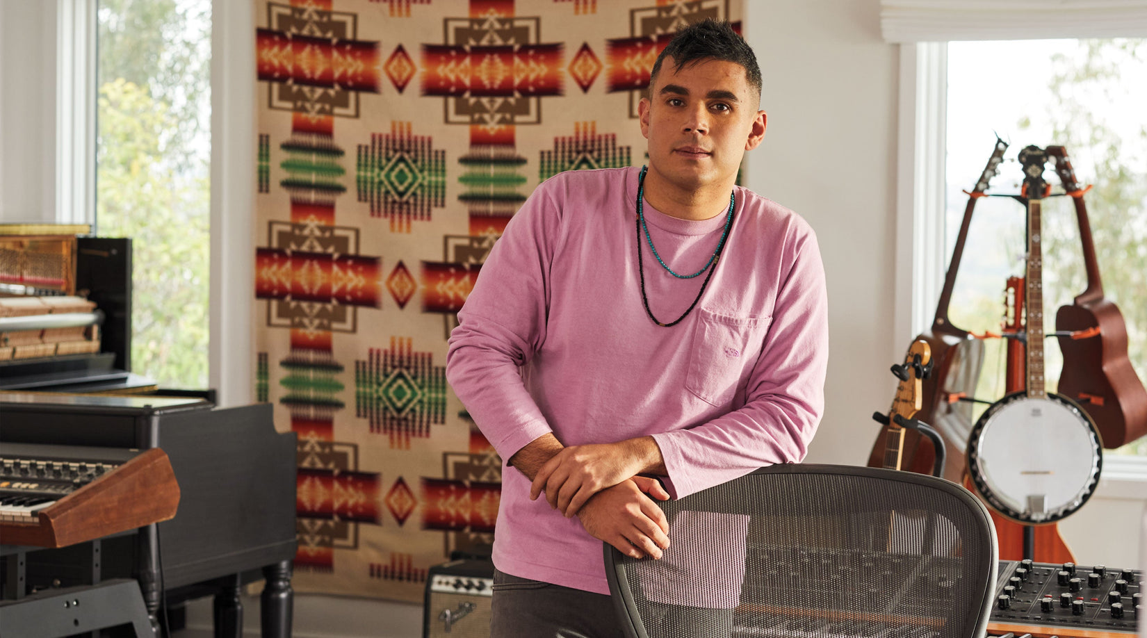 Rostam, músico, productor y compositor, de pie apoyado en una silla de oficina Aeron en grafito con instrumentos musicales al fondo.