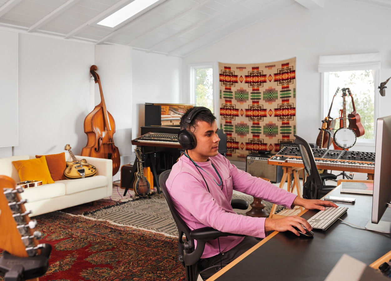 Rostam, músico, productor y compositor, sentado en una silla de oficina Aeron en grafito frente al ordenador con auriculares puestos y instrumentos musicales al fondo.